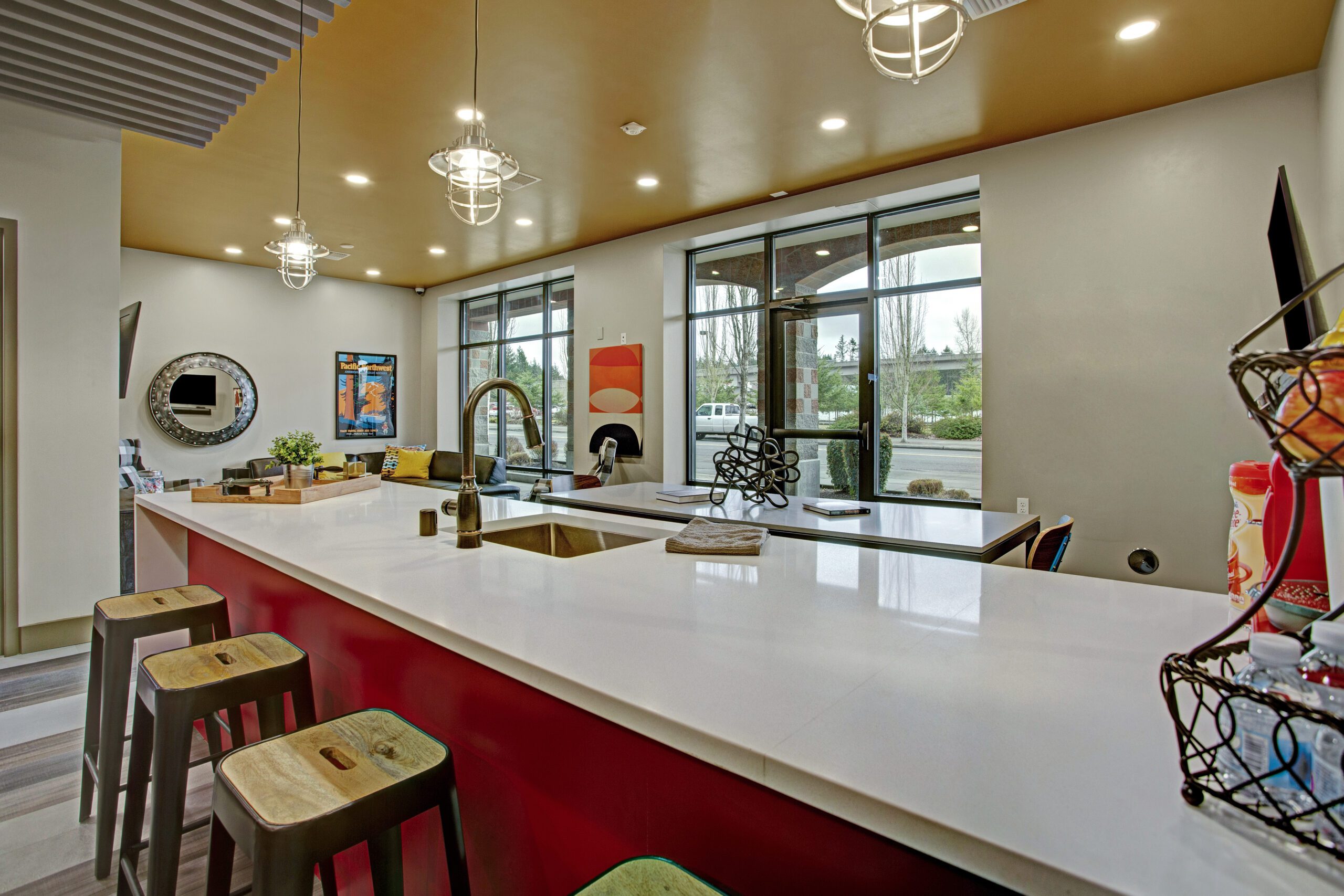 Modern kitchen with a long white island counter, four bar stools, pendant lights, and large windows. Wall art and a mirror decorate the space.